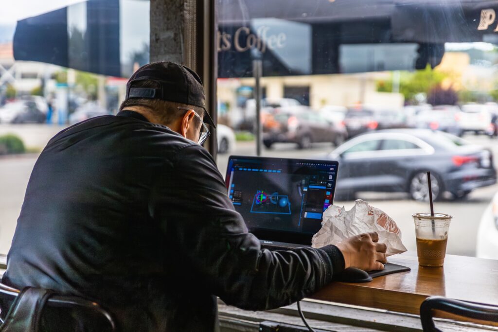 A man working on a laptop with coffee in an urban cafe, viewed from behind.