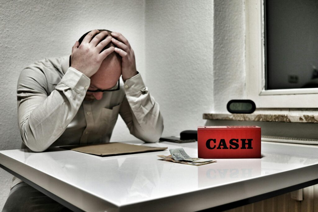 How to Stop Living Paycheck to Paycheck Bald man holding head in despair at desk with cashbox and money. Indoor office stress concept.